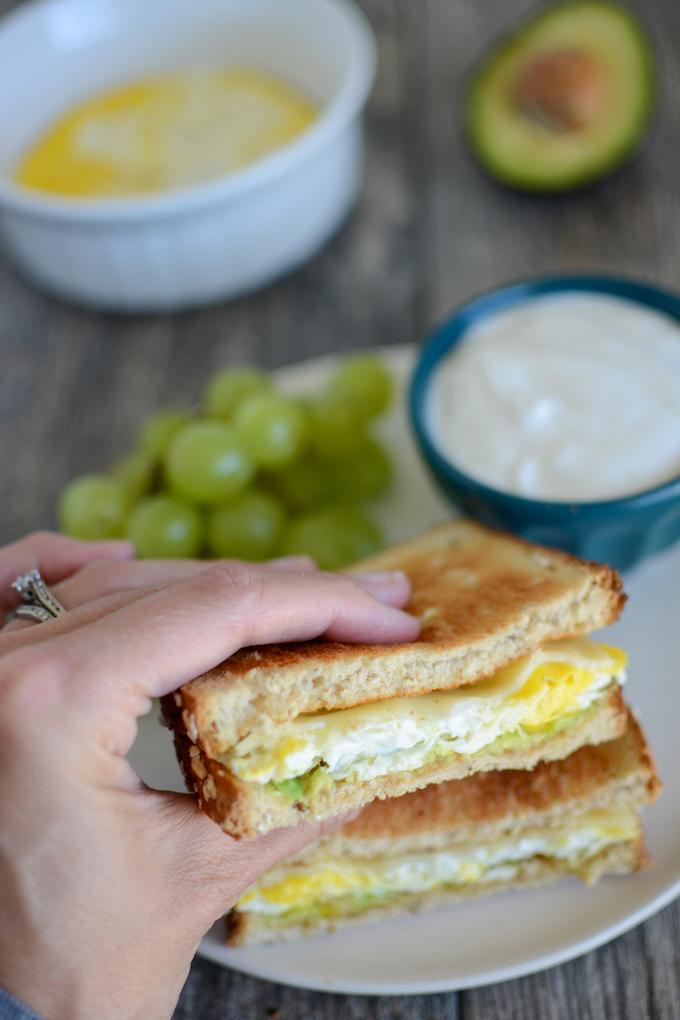 Microwave egg sandwich in hand, grapes, yogurt, egg and avocado in the background