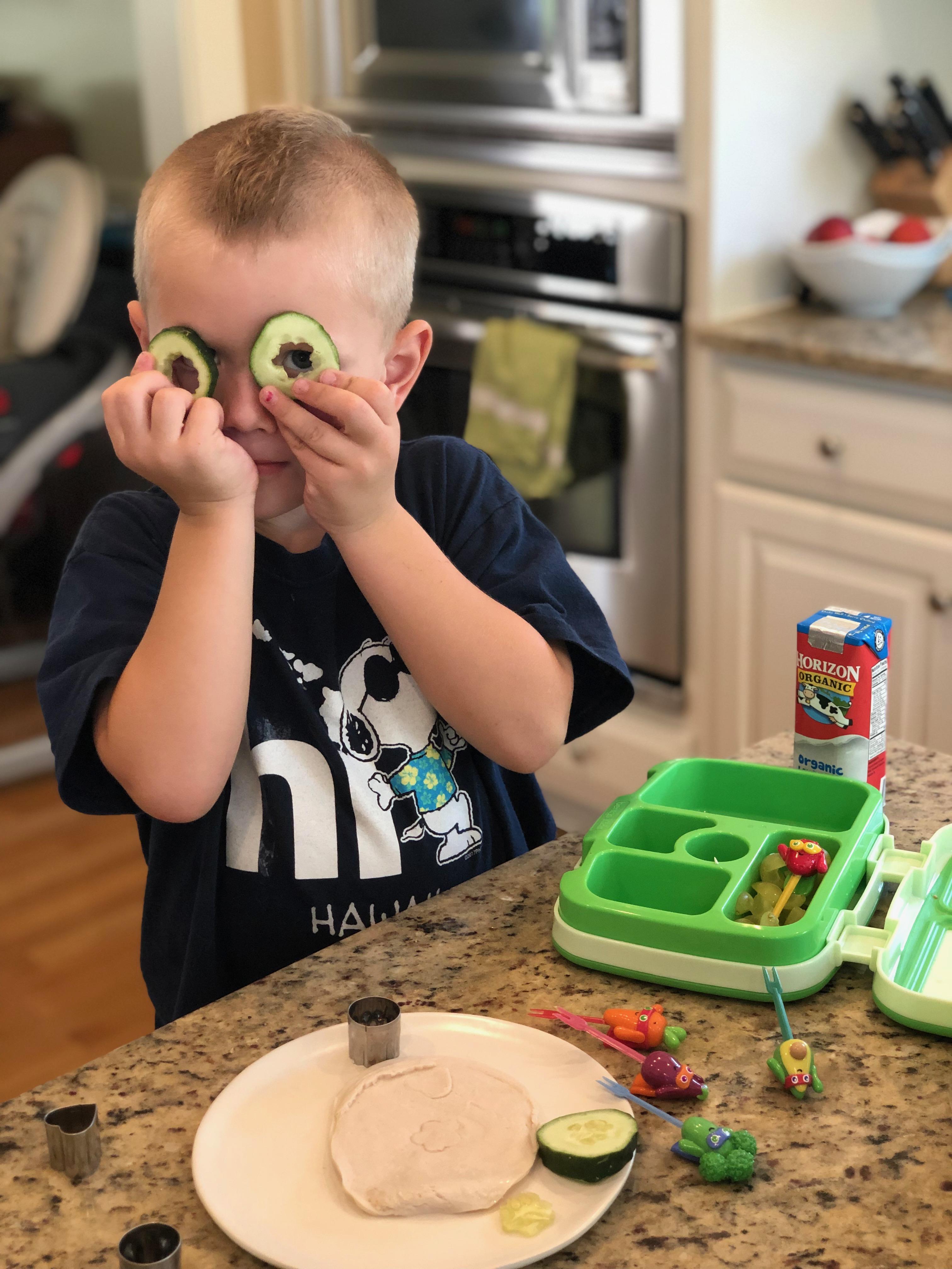 boy holding cucumber cut outs over eyes like glasses while doing school lunch packing